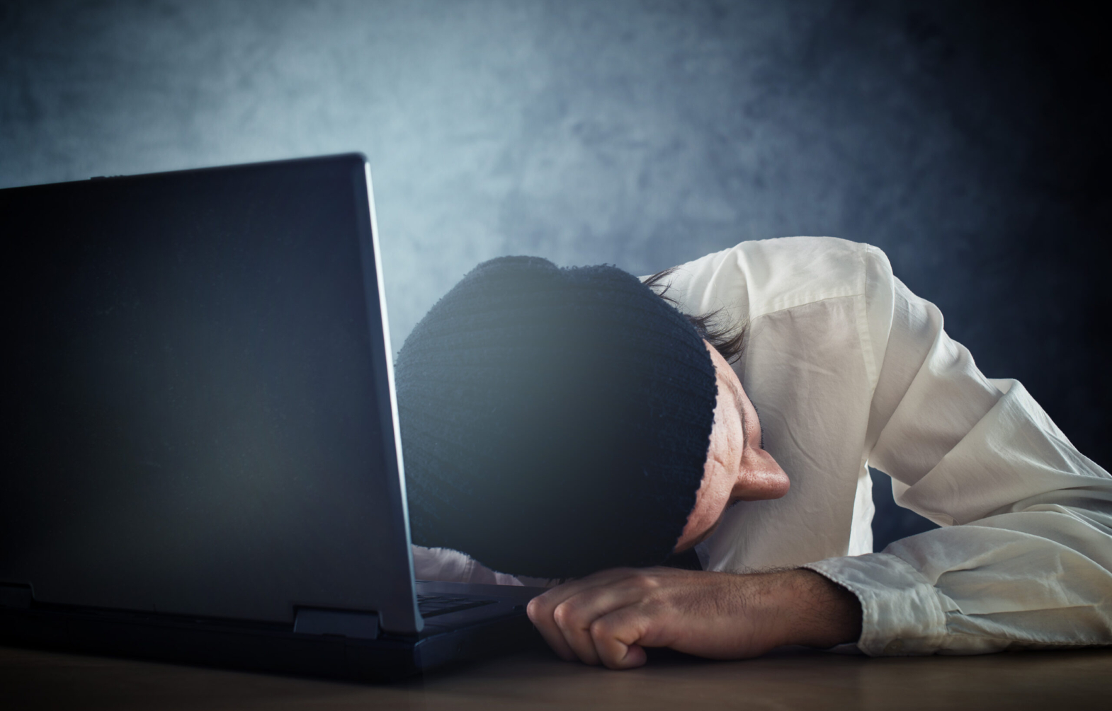 Exhausted man sleeps on laptop at office desk after working overtime.